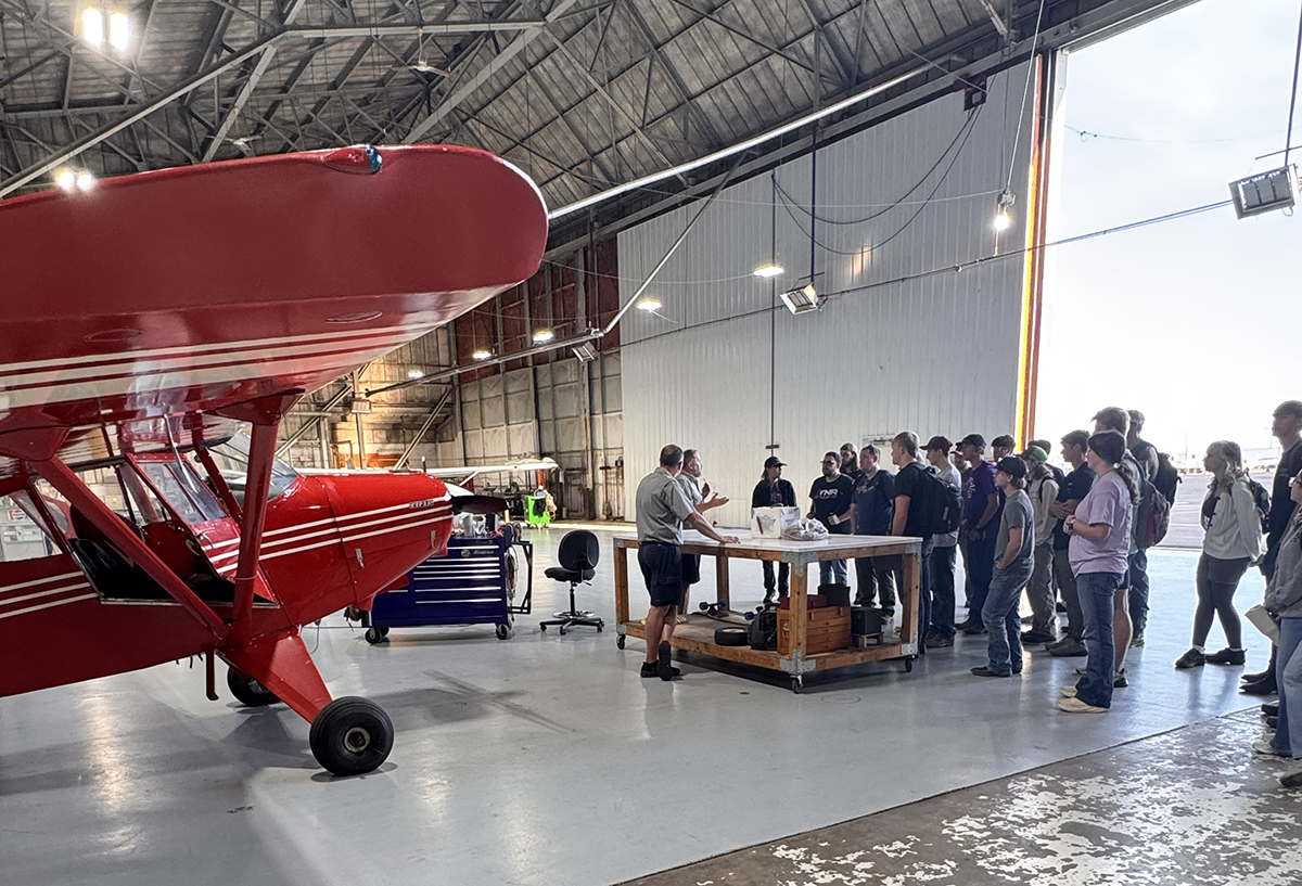 Two men address a group of students next to a red plane in a hangar.