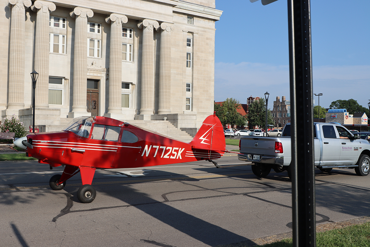 A red plane is pulled by a silver pickup truck in front of a columned building.