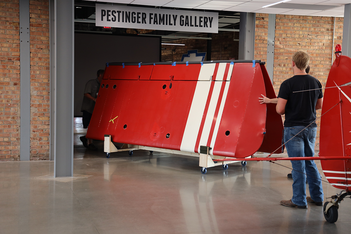 A red airplane wing is maneuvered into a room marked with a sign as Pestinger Family Gallery.