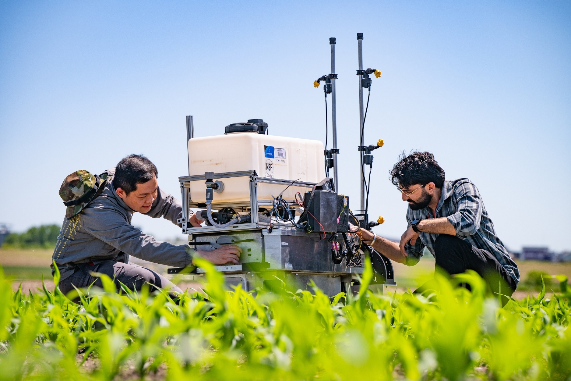 Two researchers work on agricultural equipment in a crop field, adjusting components and monitoring technology mounted on the machinery.