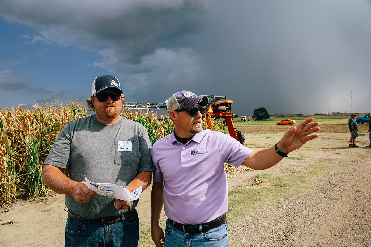 A college professor in a baseball cap has a discussion with a farmer next to a field of corn.