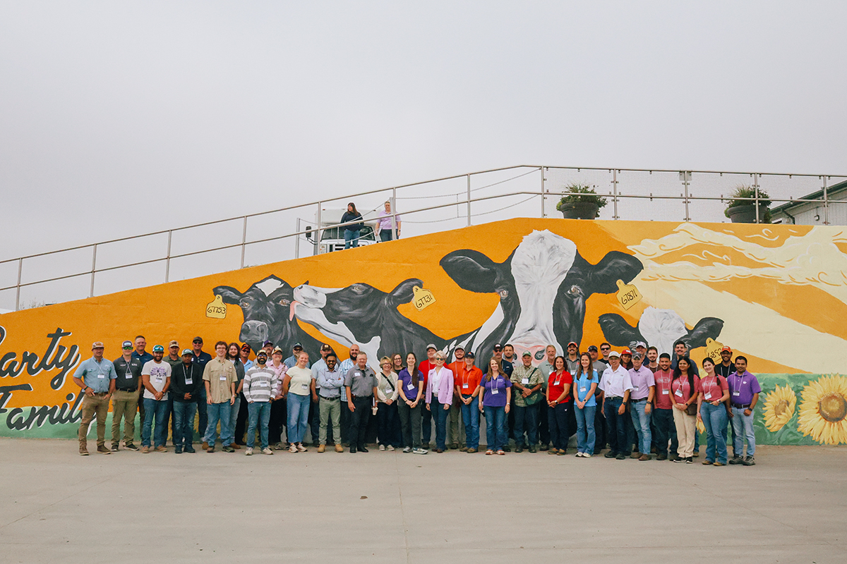 A large group of producers gathers for a portrait in front of an outdoor mural of a cow on the side of a building.