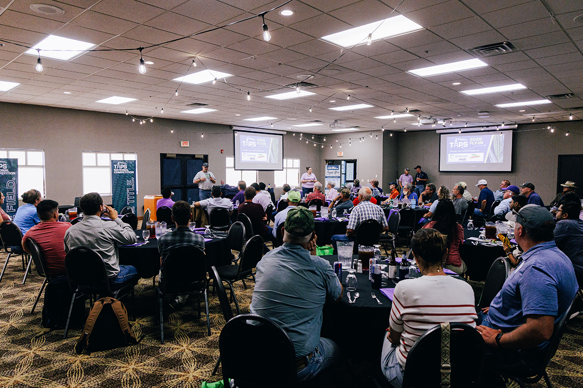 A large group of farmers listens to a presentation inside a medium-sized meeting room.