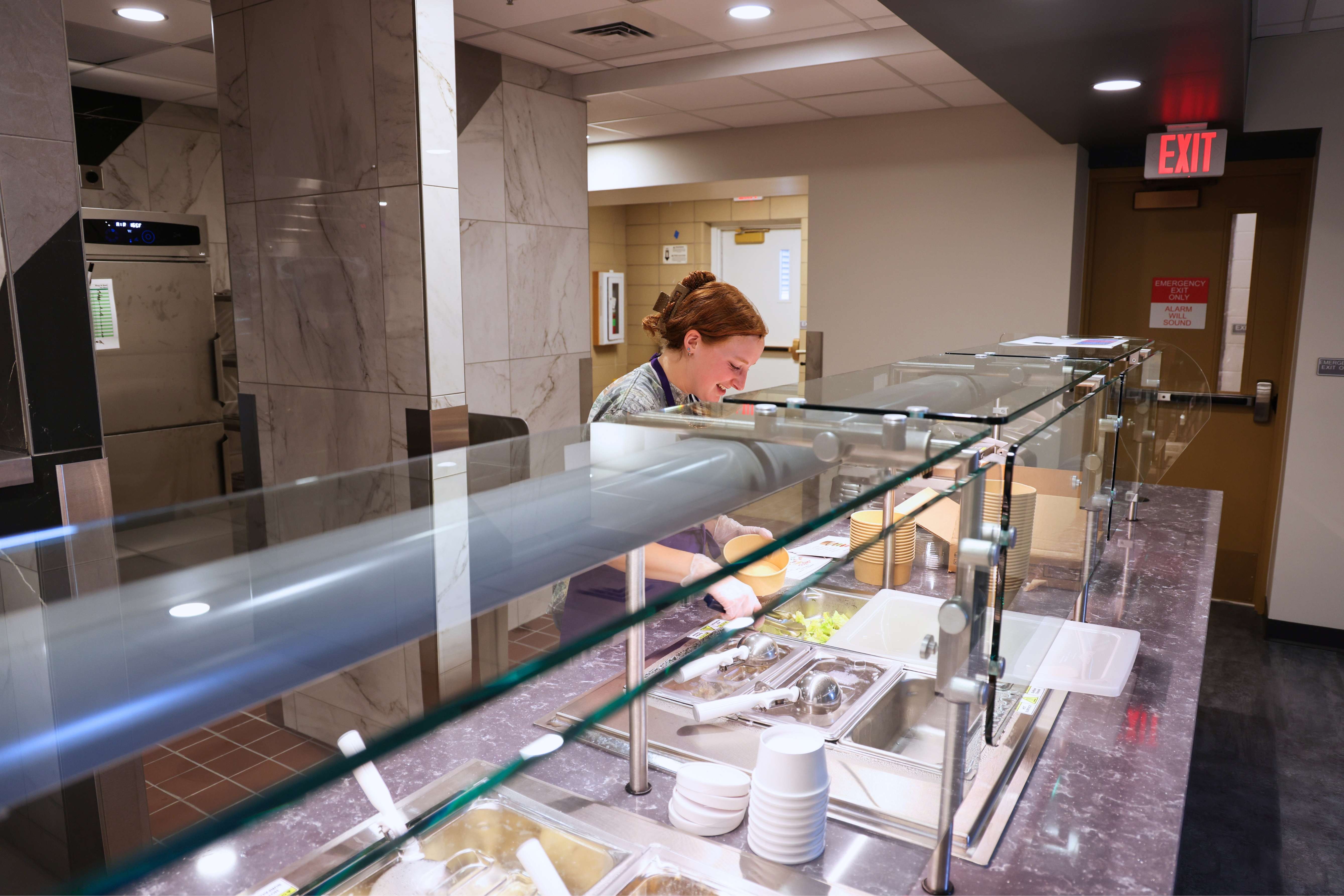 A student worker in Van Zile dining center stands behind the service counter, using tongs to put lettuce into a to-go bowl.