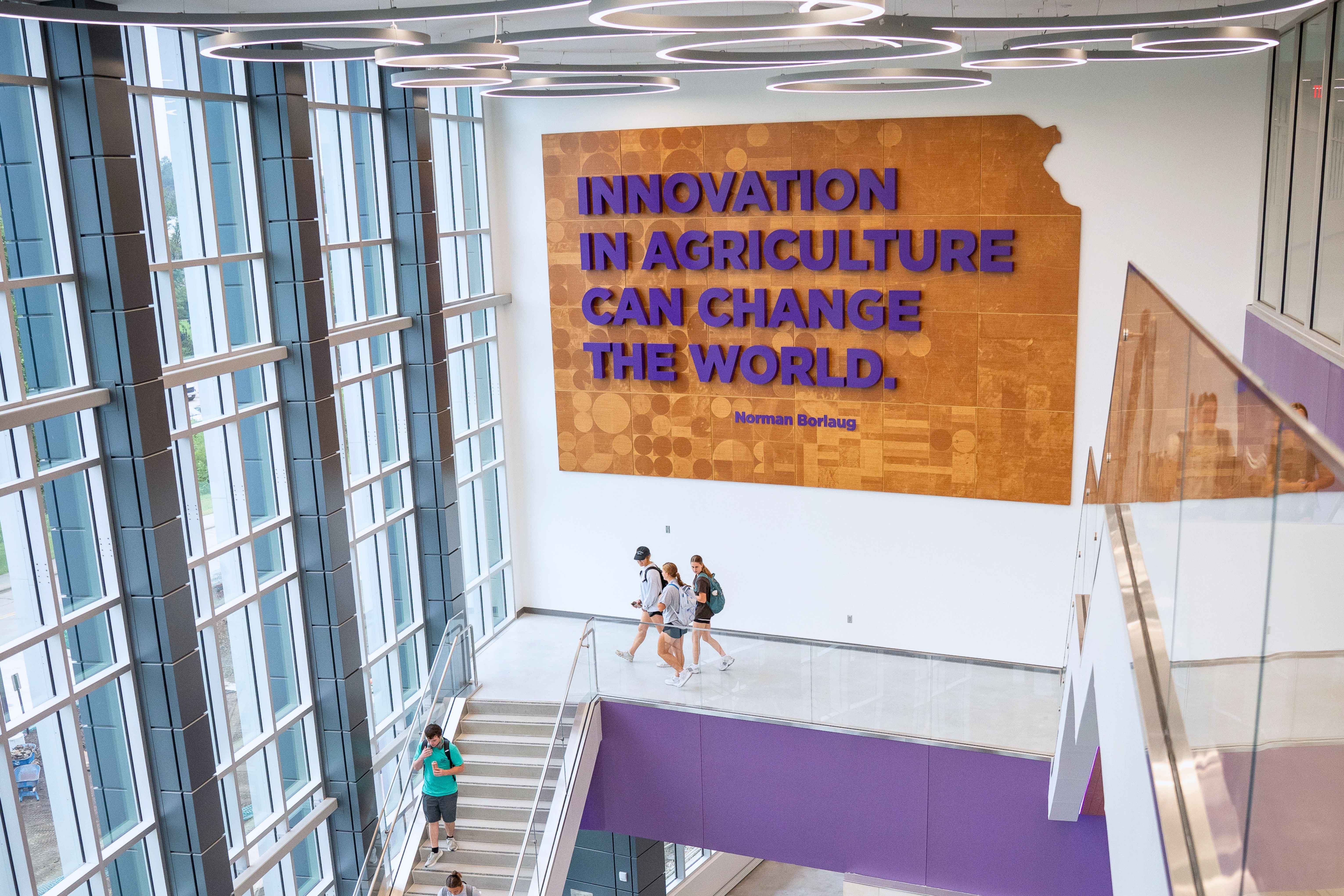 A wide shot of the interior atrium of the center, with floor-to-ceiling glass walls. On the back wall is a large wooden wall feature shaped like Kansas, with the quote: "Innovation in agriculture can change the world," Norman Borlaug, in purple block letters.
