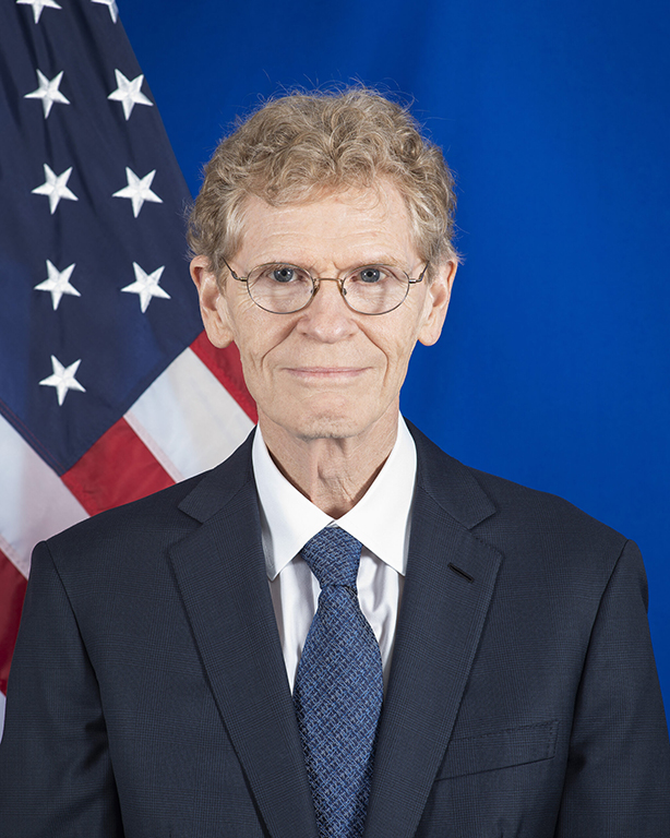 A man with circular glasses wearing a navy suit stands in front of a blue background with an American flag in the corner.