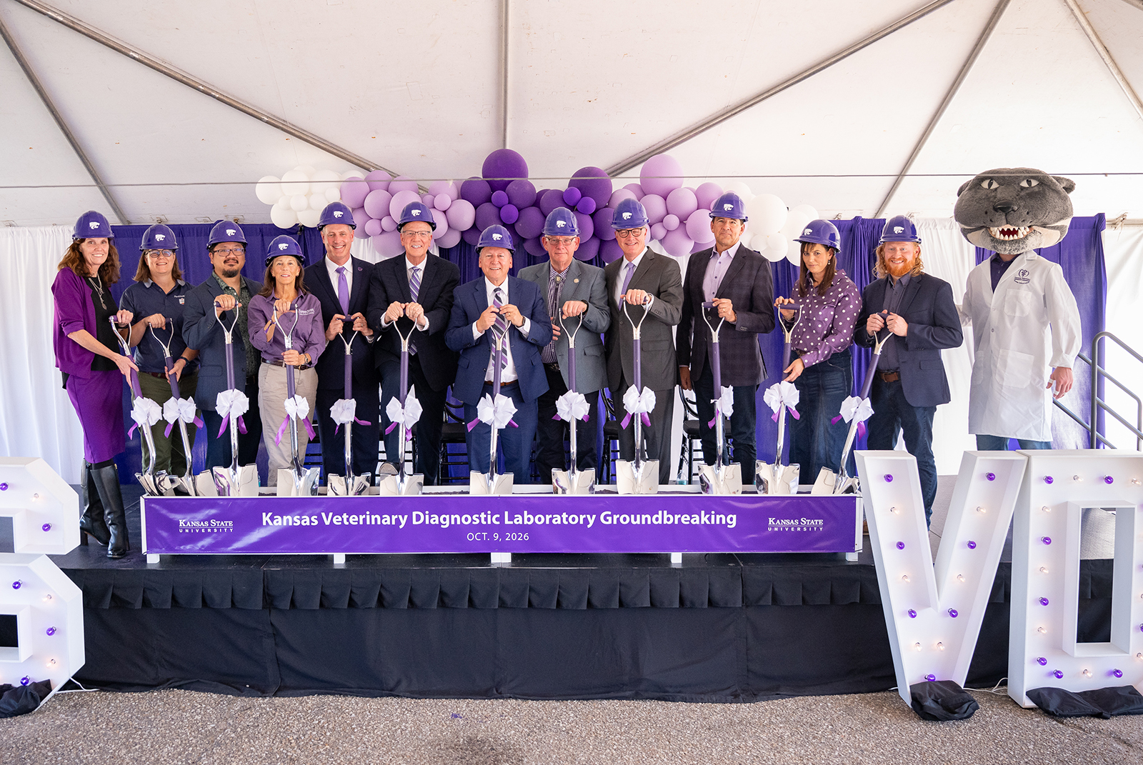 A group of officials in suits and purple hard hats hold shovels in front of a trough as part of a groundbreaking.