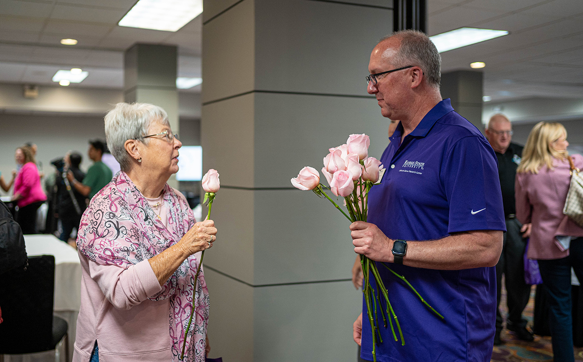 A woman in a pink outfit holding a pink rose talks to a man in a purple polo with a handful of roses.