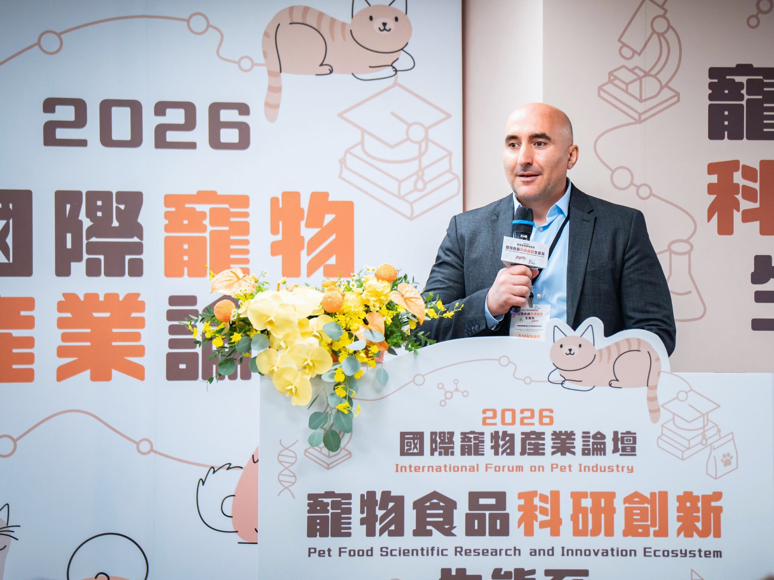 Aleksan Shanoyan stands at a podium, microphone in hand, speaking during a pet food conference