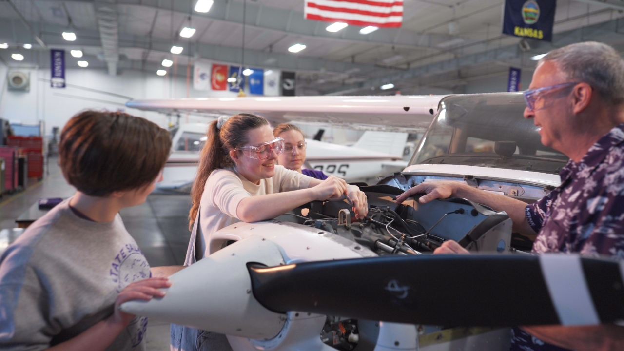 hree high school students gather around a plane with a man pointing at things in the plane's engine.