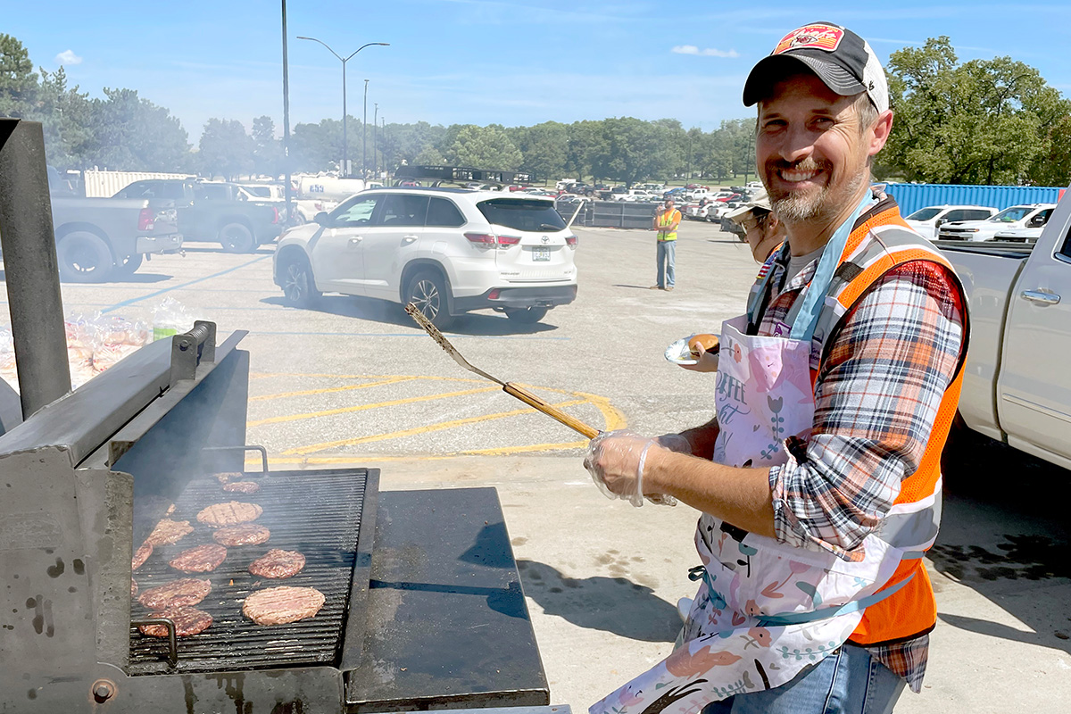 A man in a construction jacket stands at a bbq and makes burgers. 