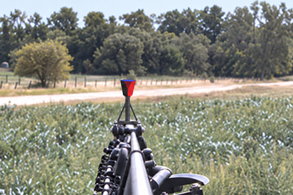 A red prismatic rectangle stands on the tip of a crop sprayer's boom over a field.