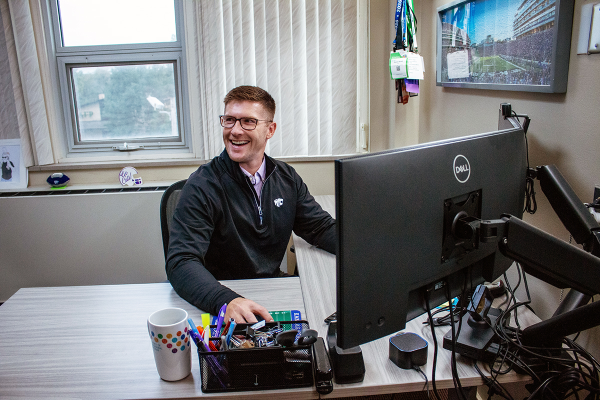 A college professor sits at a desk with his computer and smiles off camera.