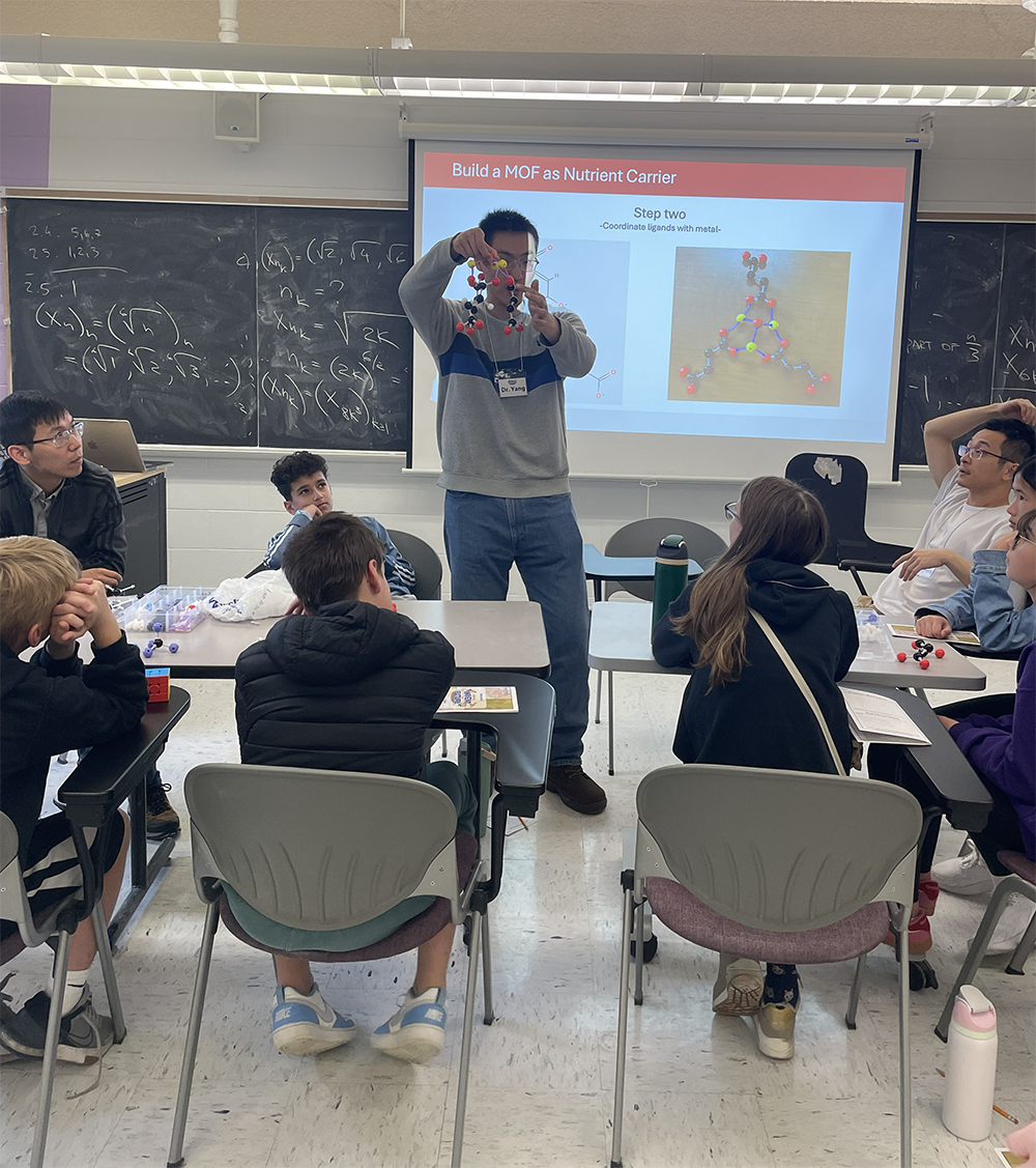 A person stands at the front of a room of middle schoolers and holds up a molecule model.