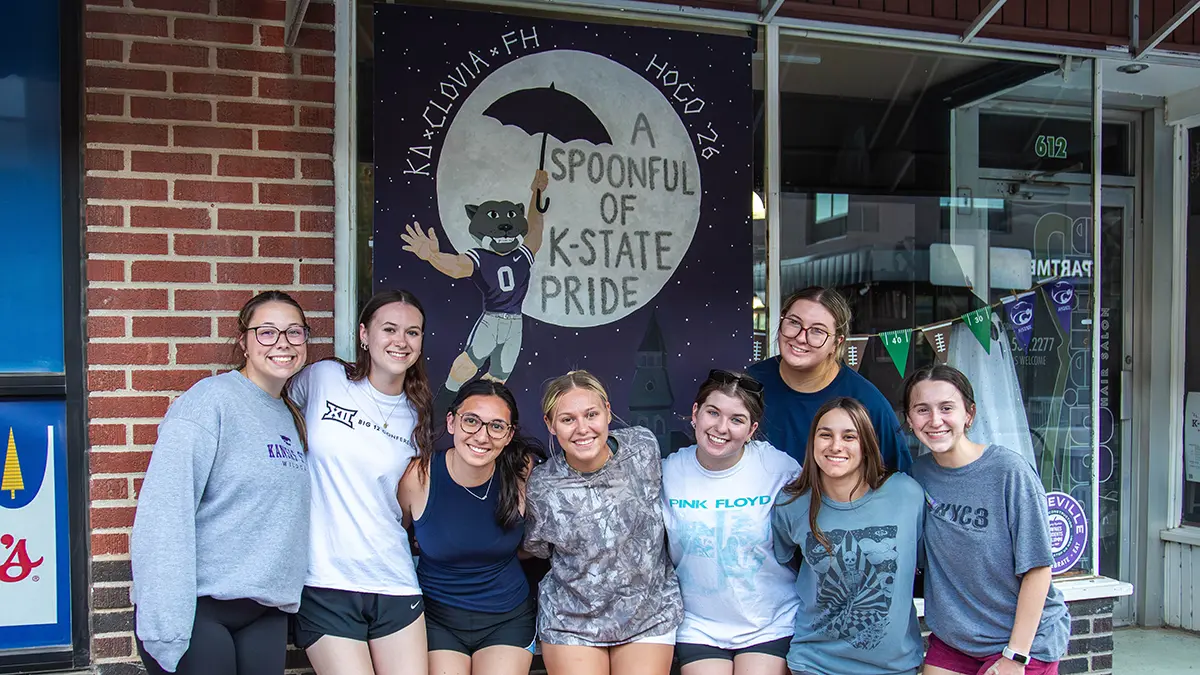 A group of sorority girls posing in front of their painting. The painint on the window is Willie the Wildcat flinging with an umbrella, with text that says ‘A spoonful of K State Pride’ inside of a moon.