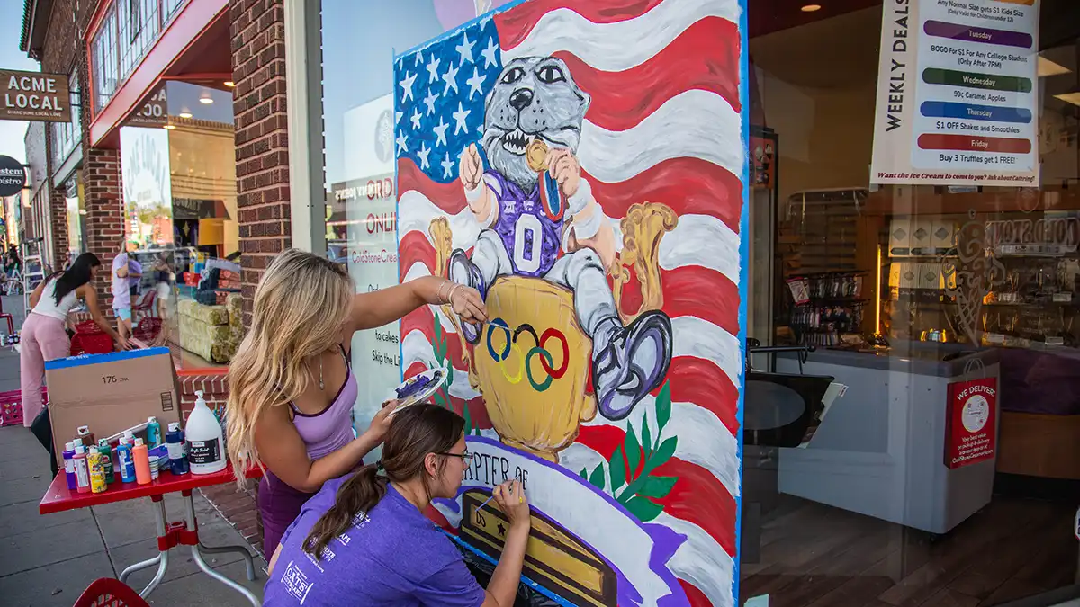 Two female student finishing the last touches of painting Willie onto a golden trophie with the American Flag behind him.