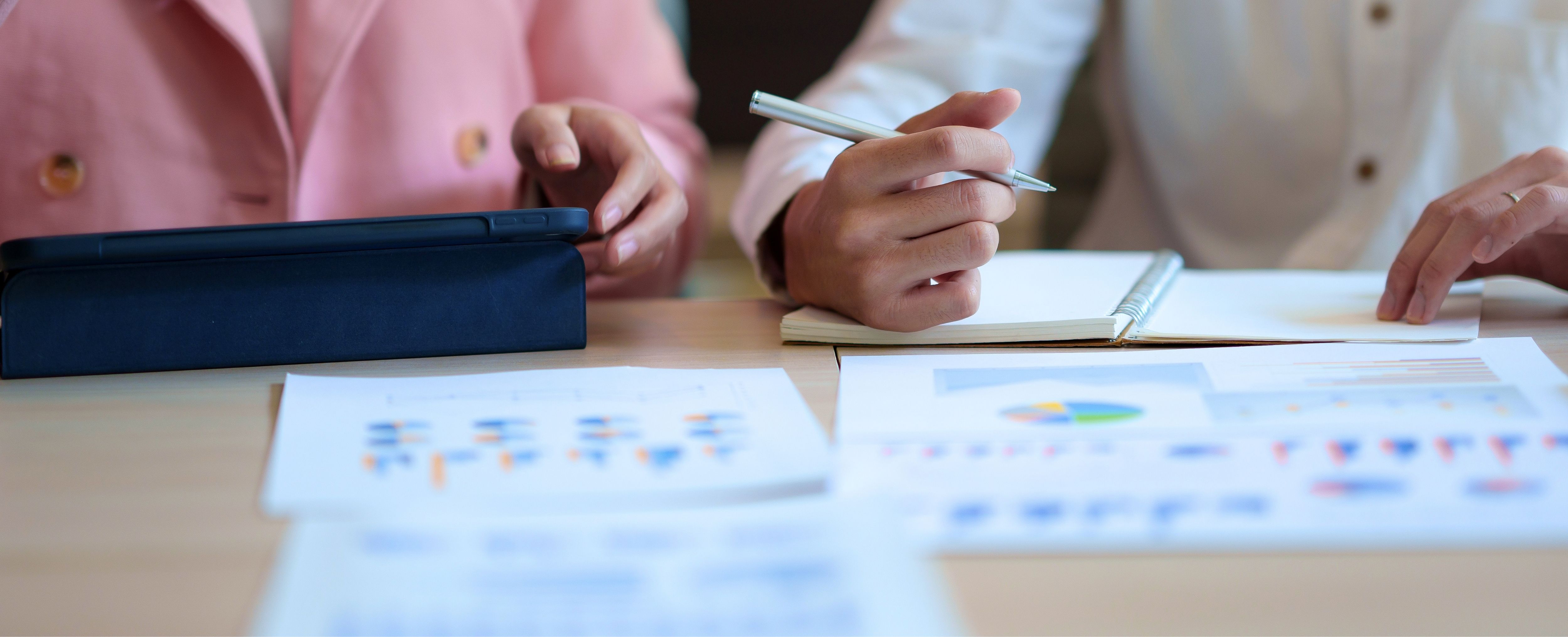 Two people discussing charts at a table. One person holds a tablet, while one is writing in a notebook.