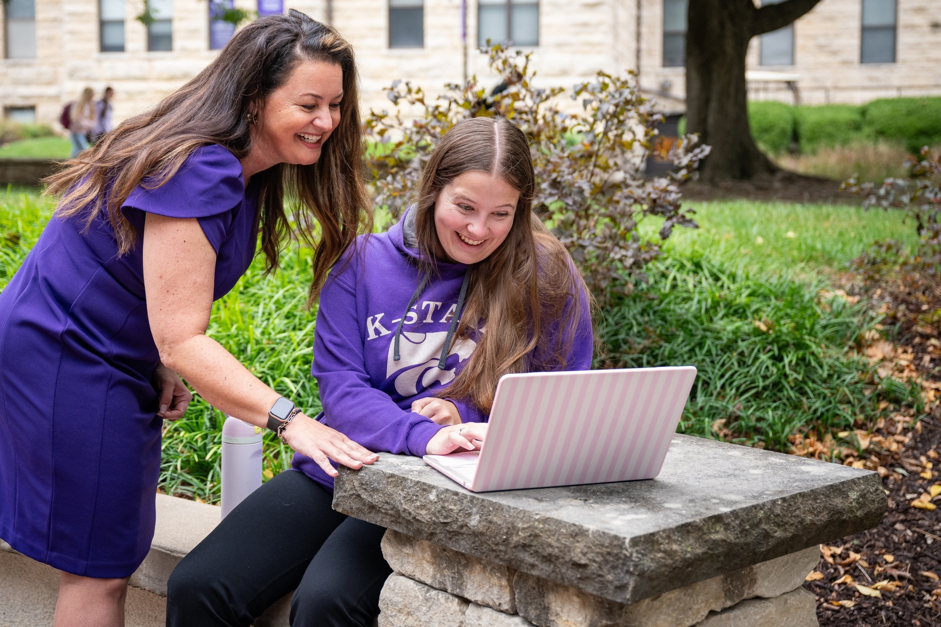 Jessica Lane stands talking to a student outside of Bluemont Hall. The student sits with their computer on a limestone post while showing Lane something on their screen.