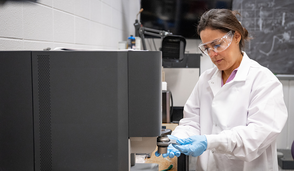 A woman in a white lab coat adjusts a piece of equipment with a white board in the background.