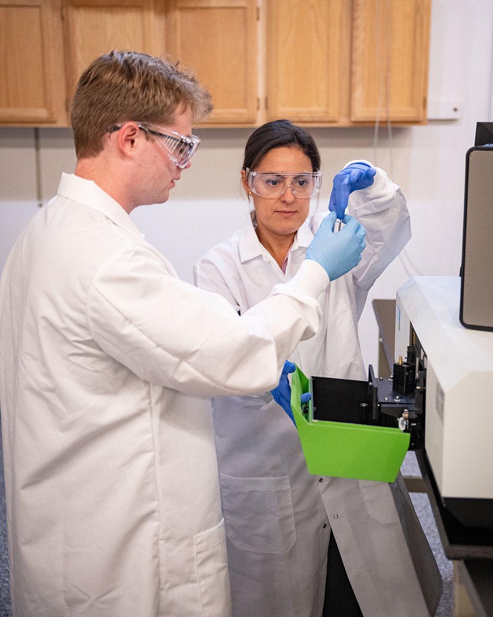 A woman and young man wearing lab coats examine a machine in a room with wooden cabinets.