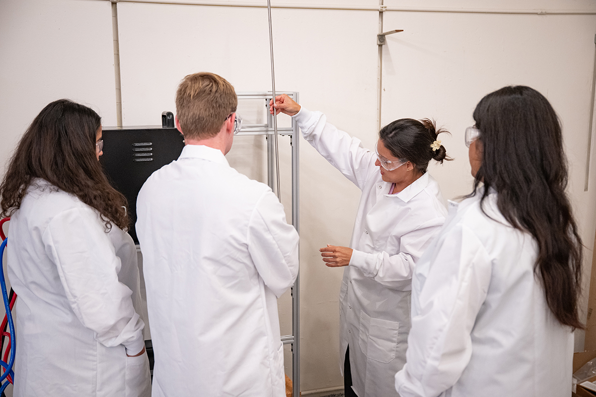Four people in white lab coats stand with their backs to the camera examining a metal tube.