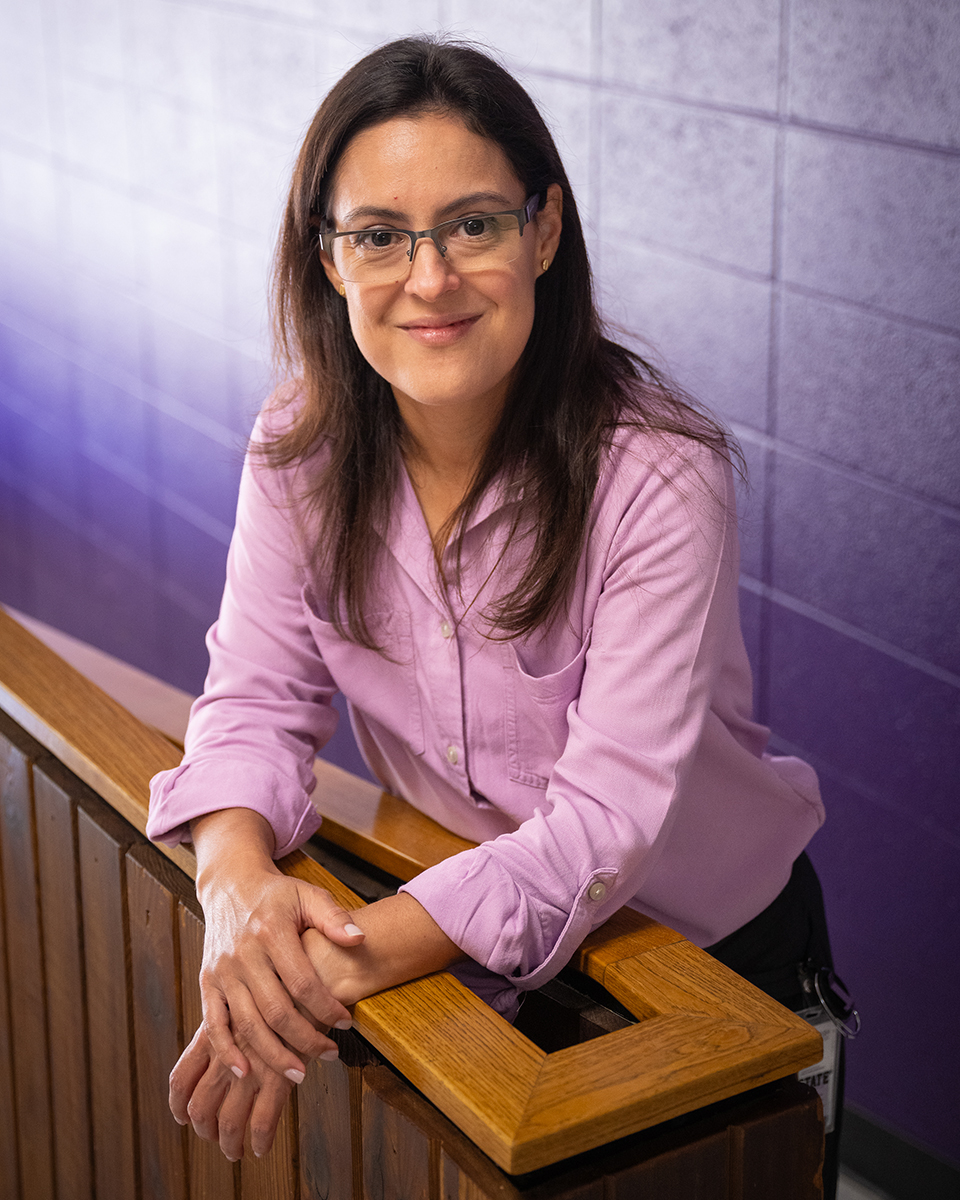 A vertical portrait shows a woman in a pink long-sleeved shirt leaning against a wooden bannister in front of a purple wall.