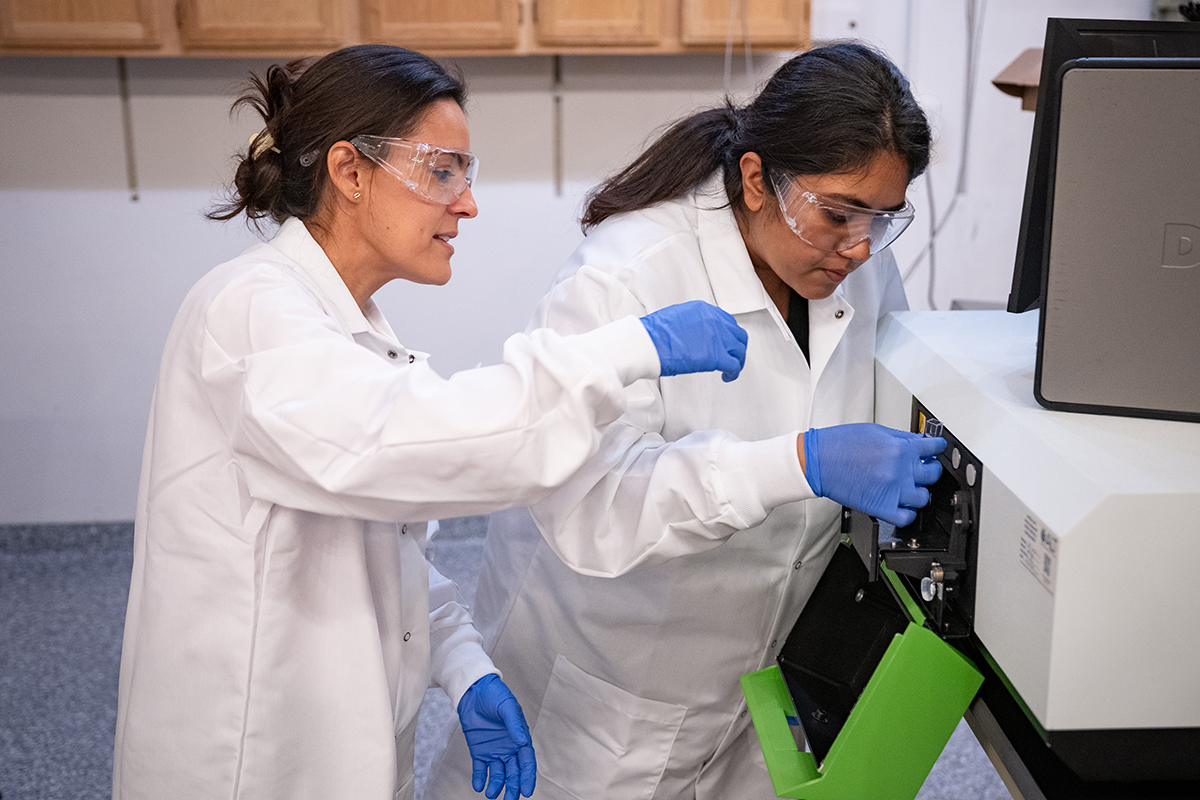 Two women in white lab coats and blue safety globes examine a machine with a lime green drawer open.