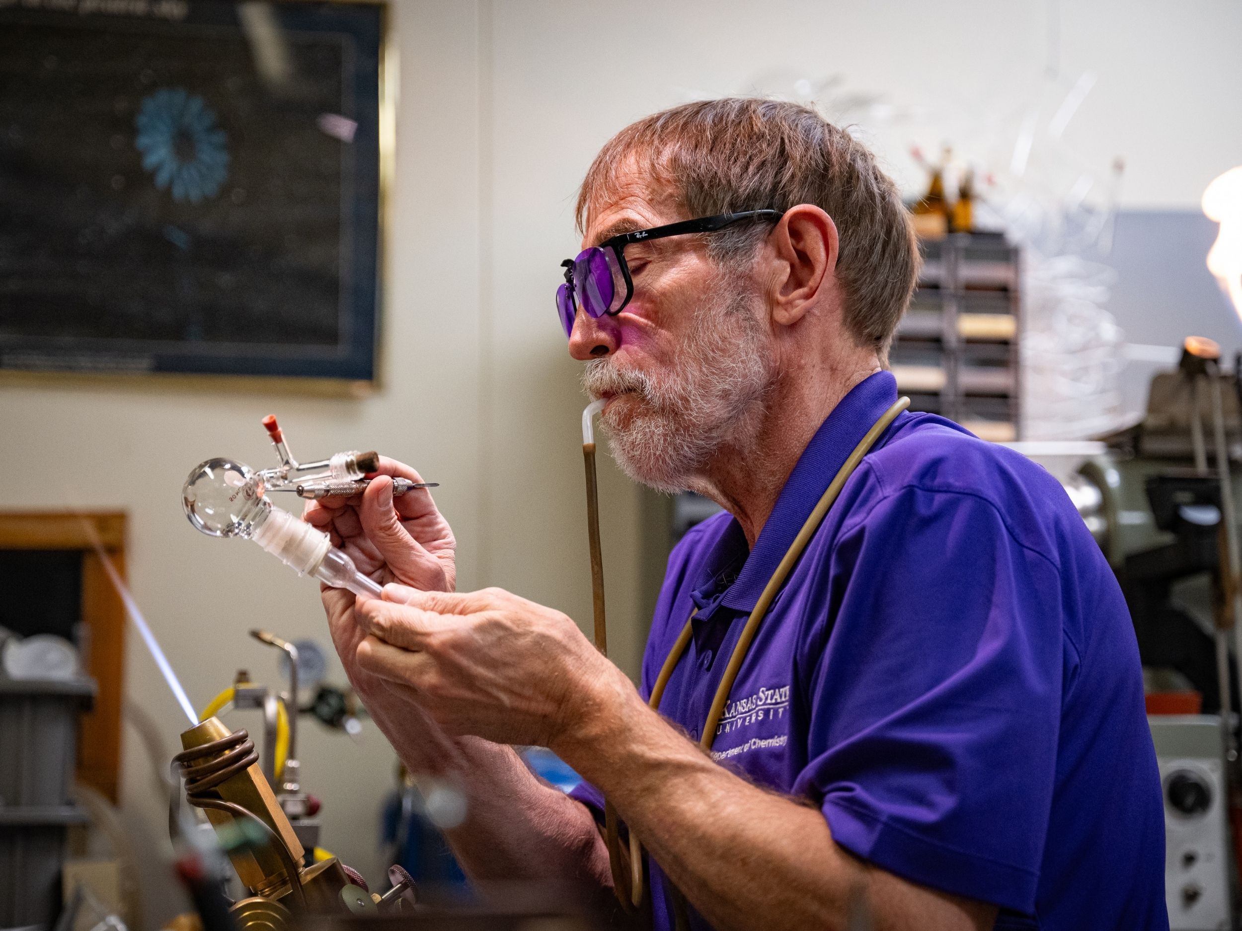 Jim Hodgson sits at his bench, using a blow hose to sustain pressure in a glass apparatus while shaping it with tweezers.