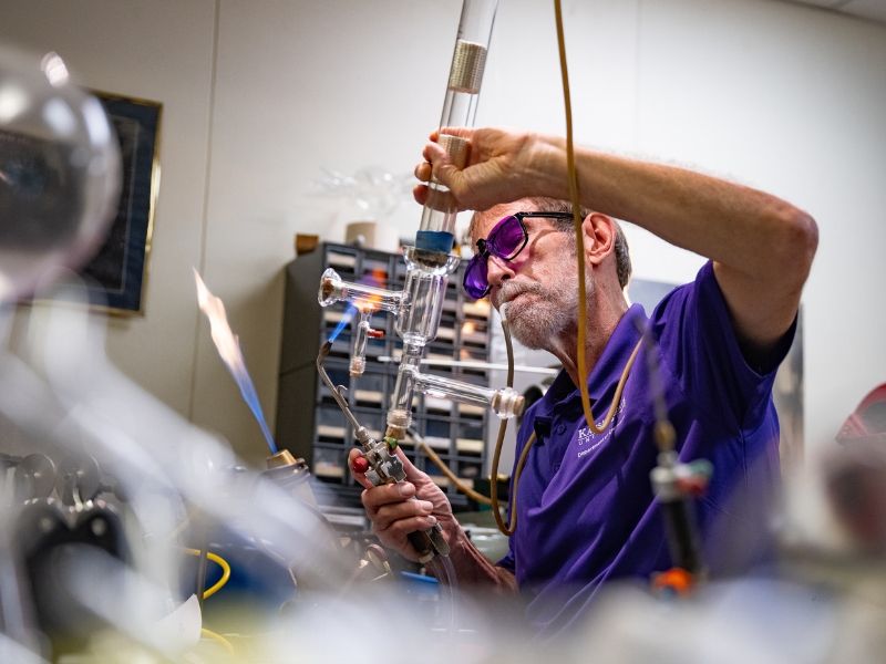 Jim Hodgson sits at his workbench, heating a piece of glass tubing. He holds a blow hose in his mouth to maintain pressure within the softening glass.