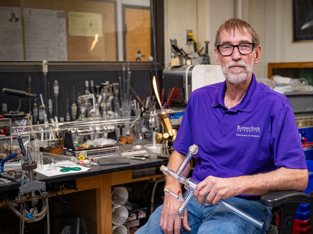 Jim Hodgson sits at his workbench, smiling while holding a piece of glass tubing.