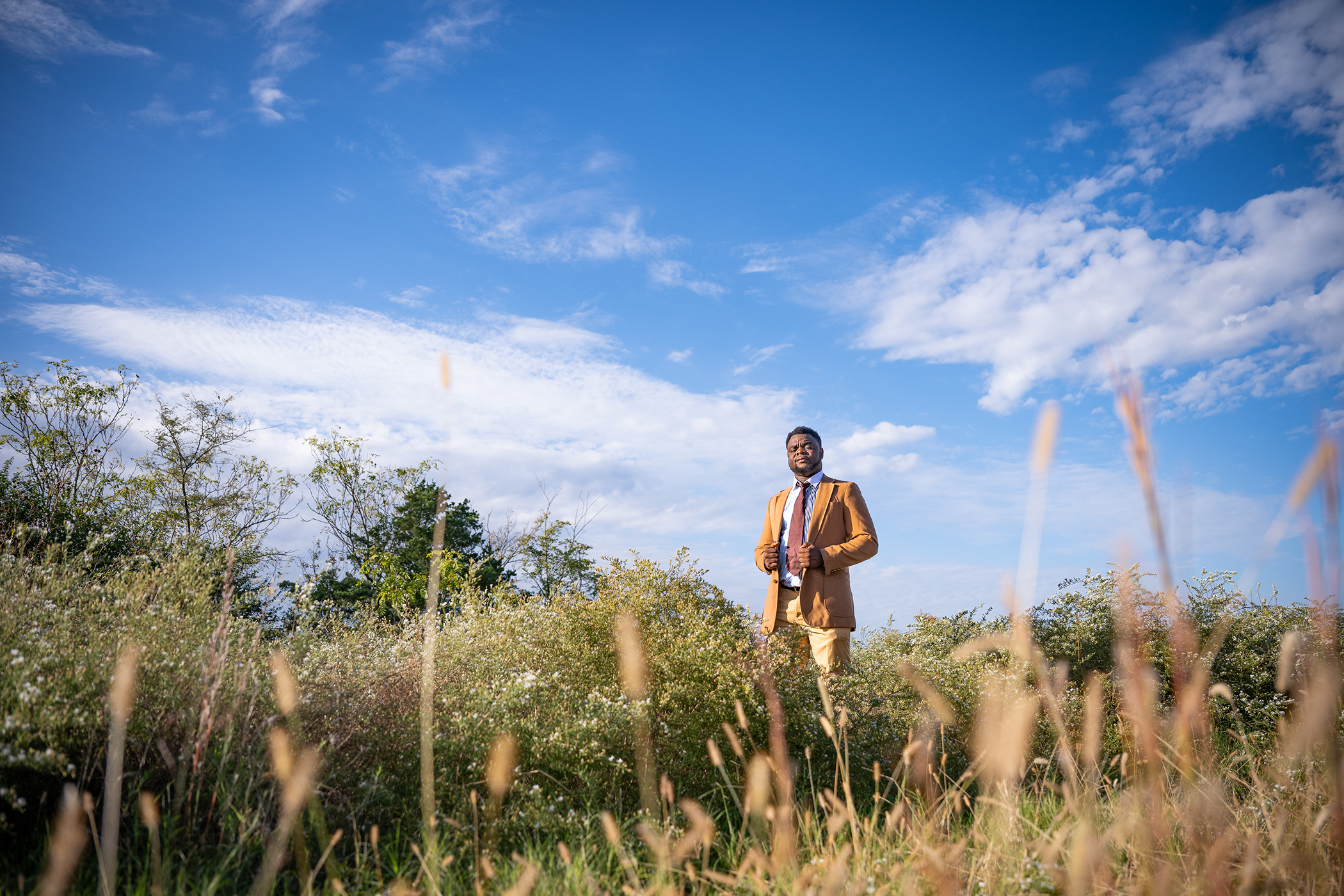 A researcher in a brown sportcoat stands in the middle of a grassy prairie.