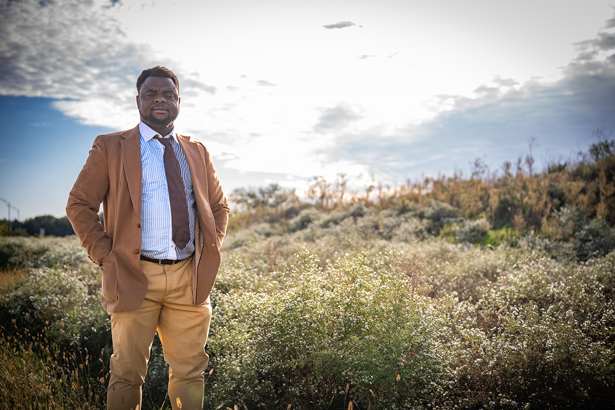 A researcher in a brown sportcoat stands in a prairie with his hands in his pockets.