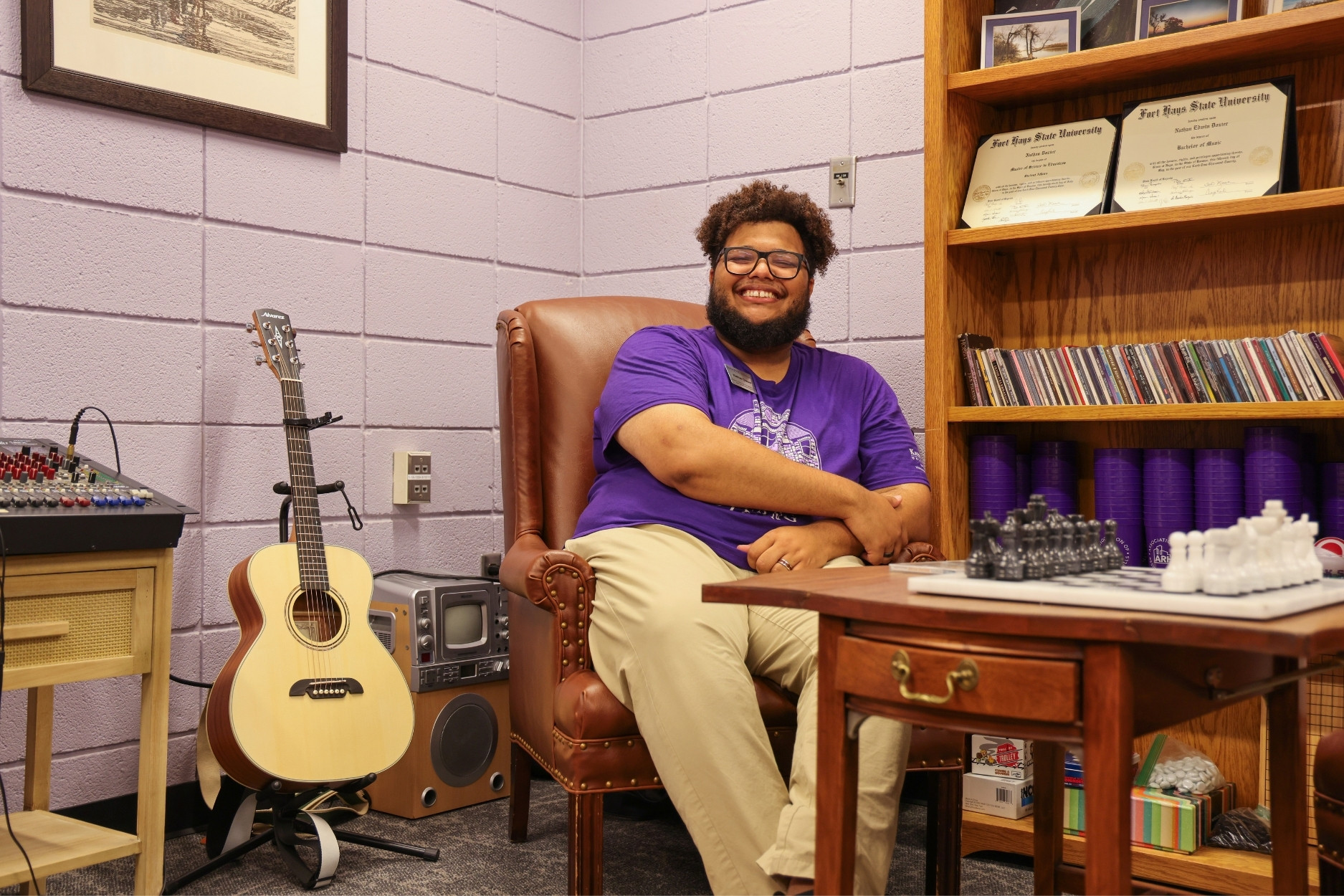 Nathan Dozier sits in an office. In the foreground is a table with a chess set. In the background sits a bookshelf, a guitar, and a sound amplifier.