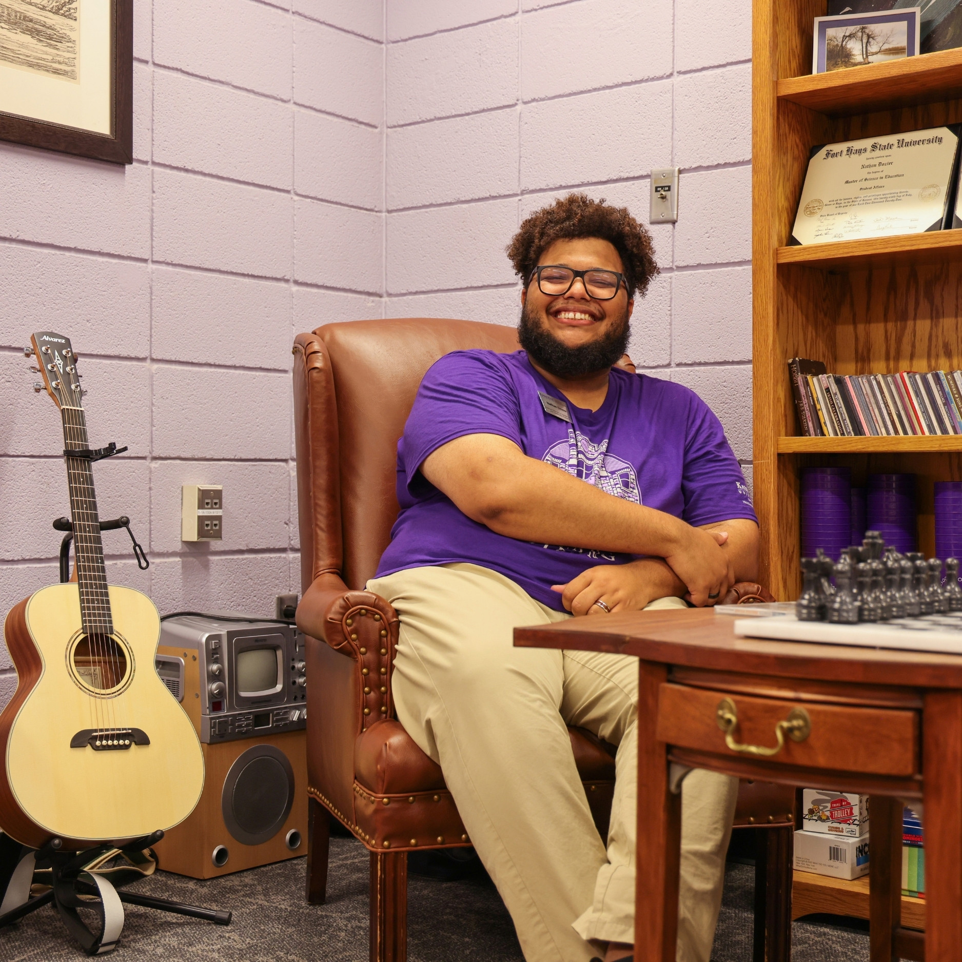 Nathan Dozier sits in an office. In the foreground is a table with a chess set. In the background sits a bookshelf, a guitar, and a sound amplifier.