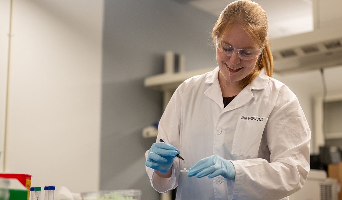 A student wearing a lab coat and safety glasses holds up a slide.