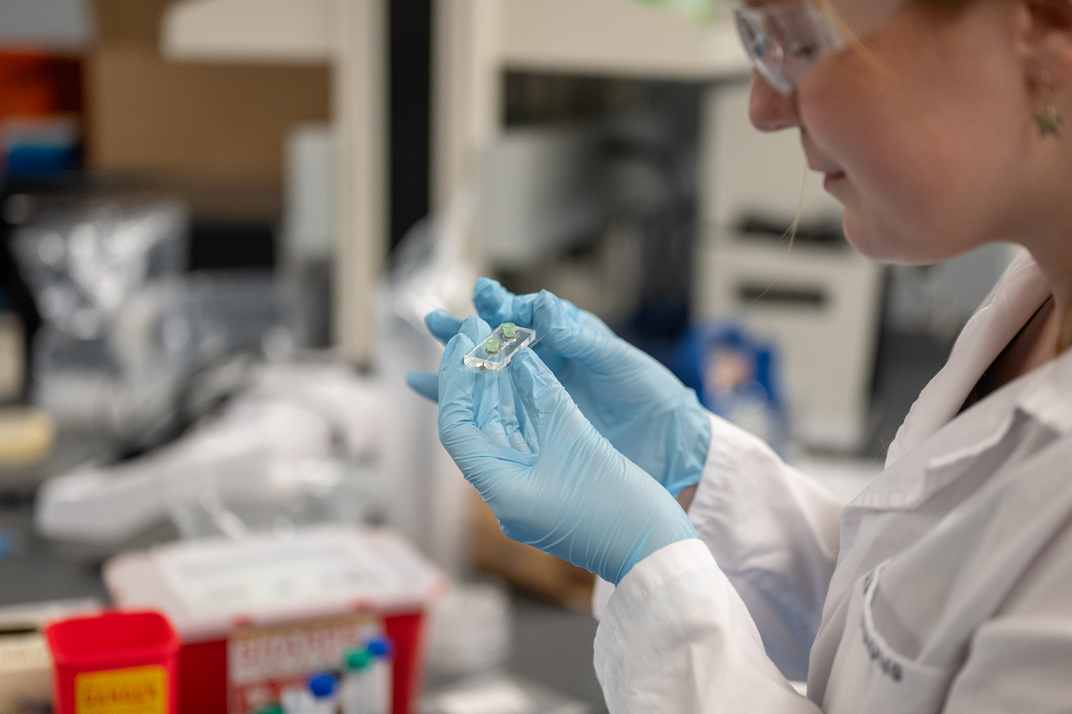 A female undergraduate student in a lab coat and safety glasses works with a small chip under a hood.