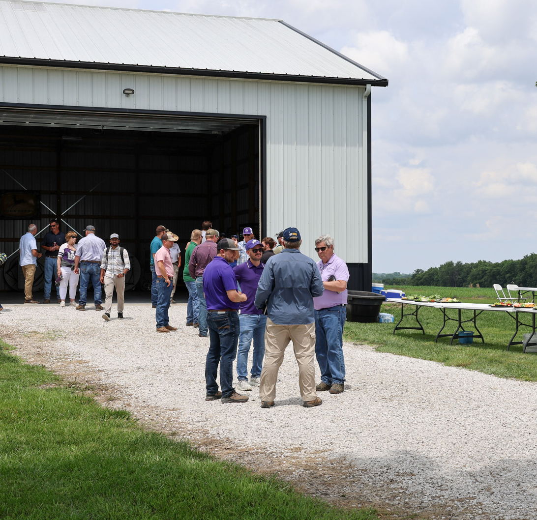 A group of people stands outside a white barn with tables of food and coffee set up nearby