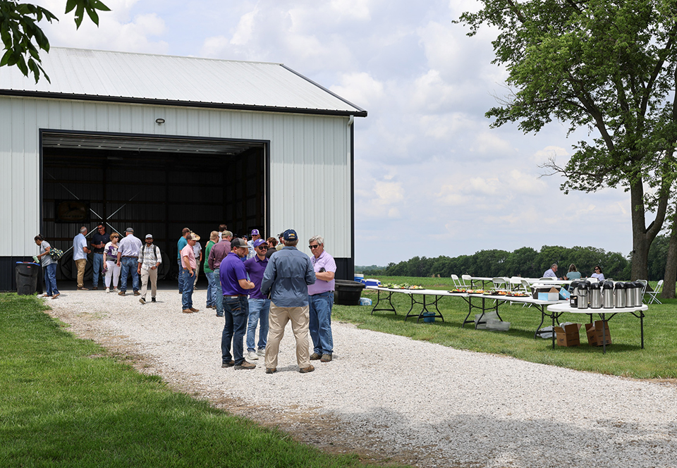 A group of people stands outside a white barn with tables of food and coffee set up nearby.