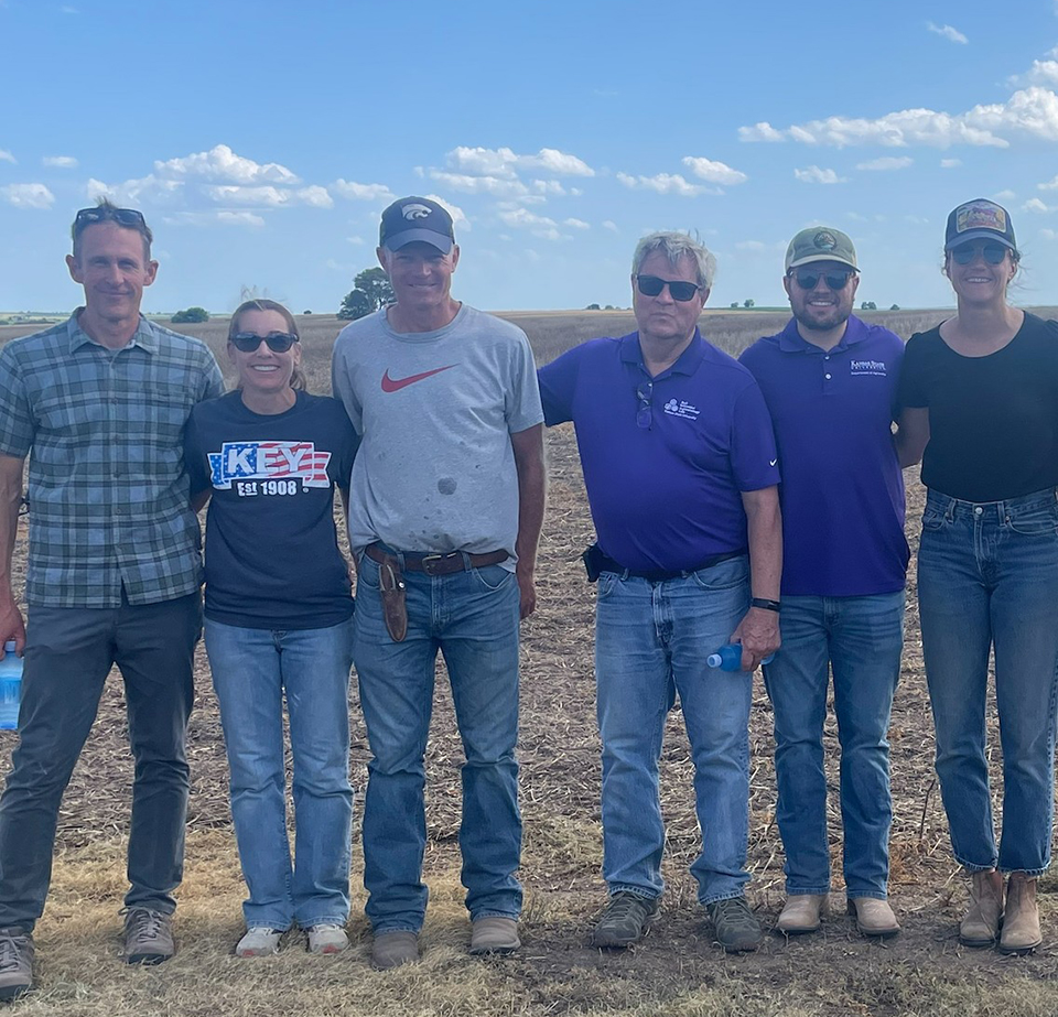 A group of six people stand in a line outside in a field with a blue sky behind them.