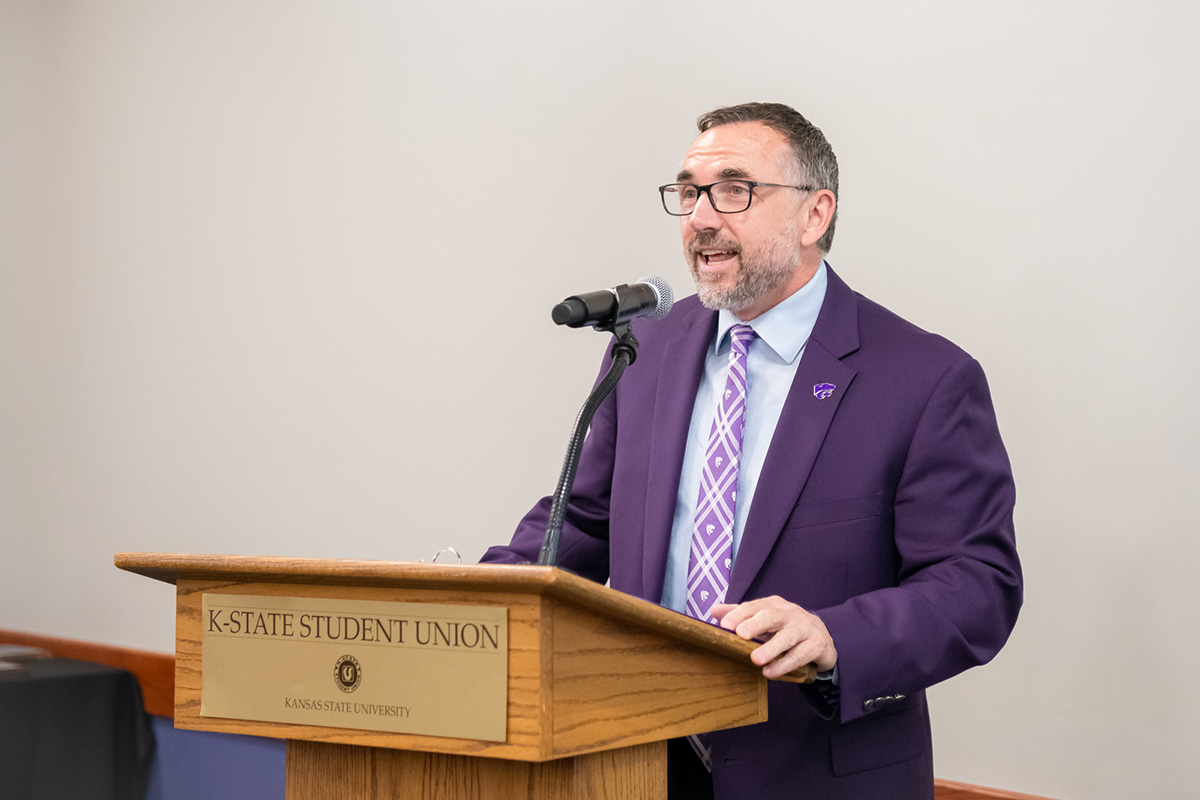 A man wearing a dark purple blazer, glasses and a purple pattern tie speaks at a wooden podium with a microphone.