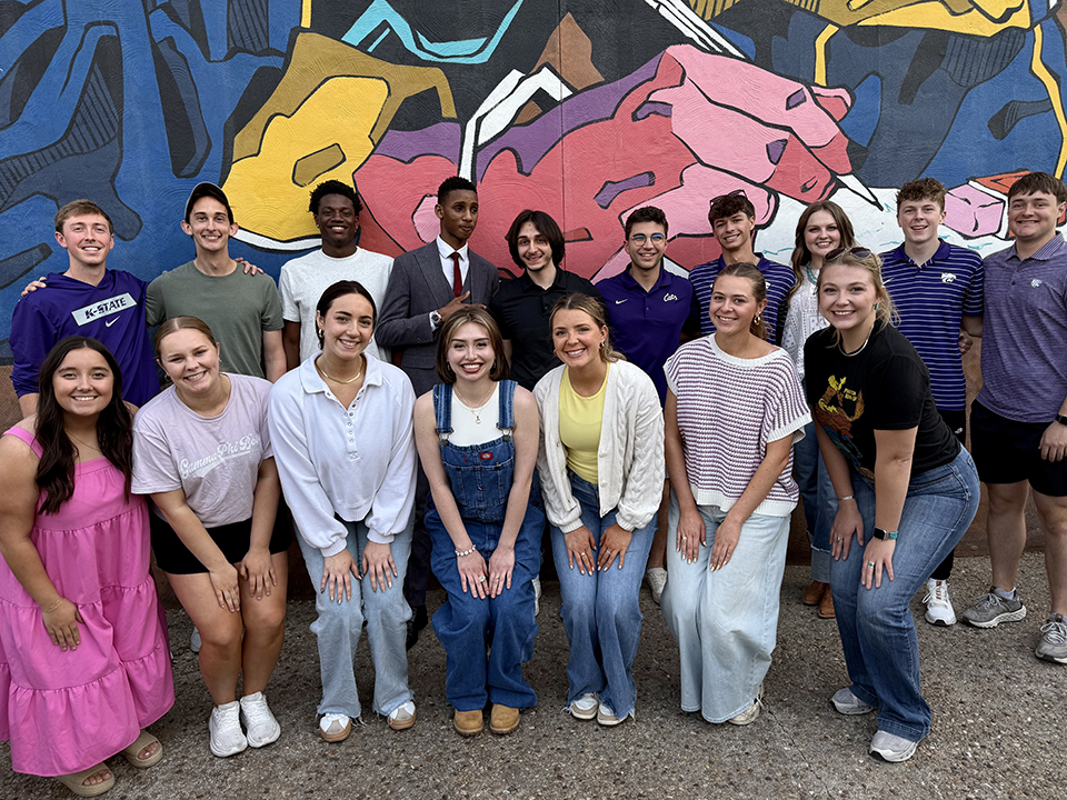 A group of several college-aged students pose for a group photo in front of a brightly colored outdoor mural.