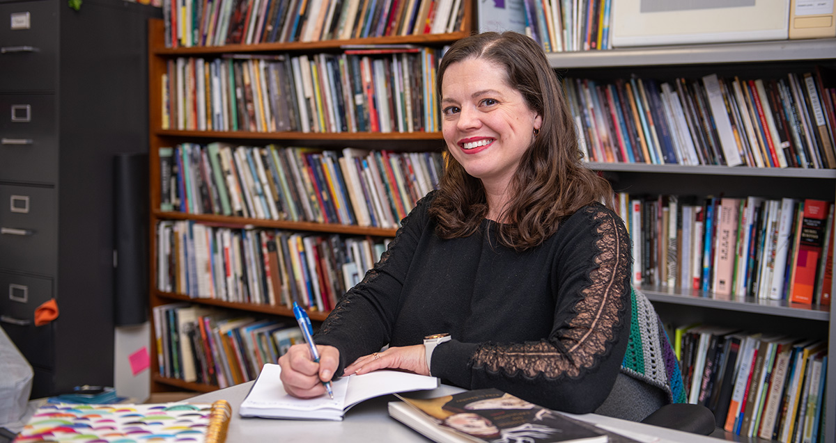 Woman smiles for a portrait while sitting at a table with a book and pen.