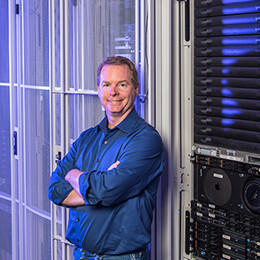 A man in a blue button-up shirt crosses his arms and smiles for a photo.