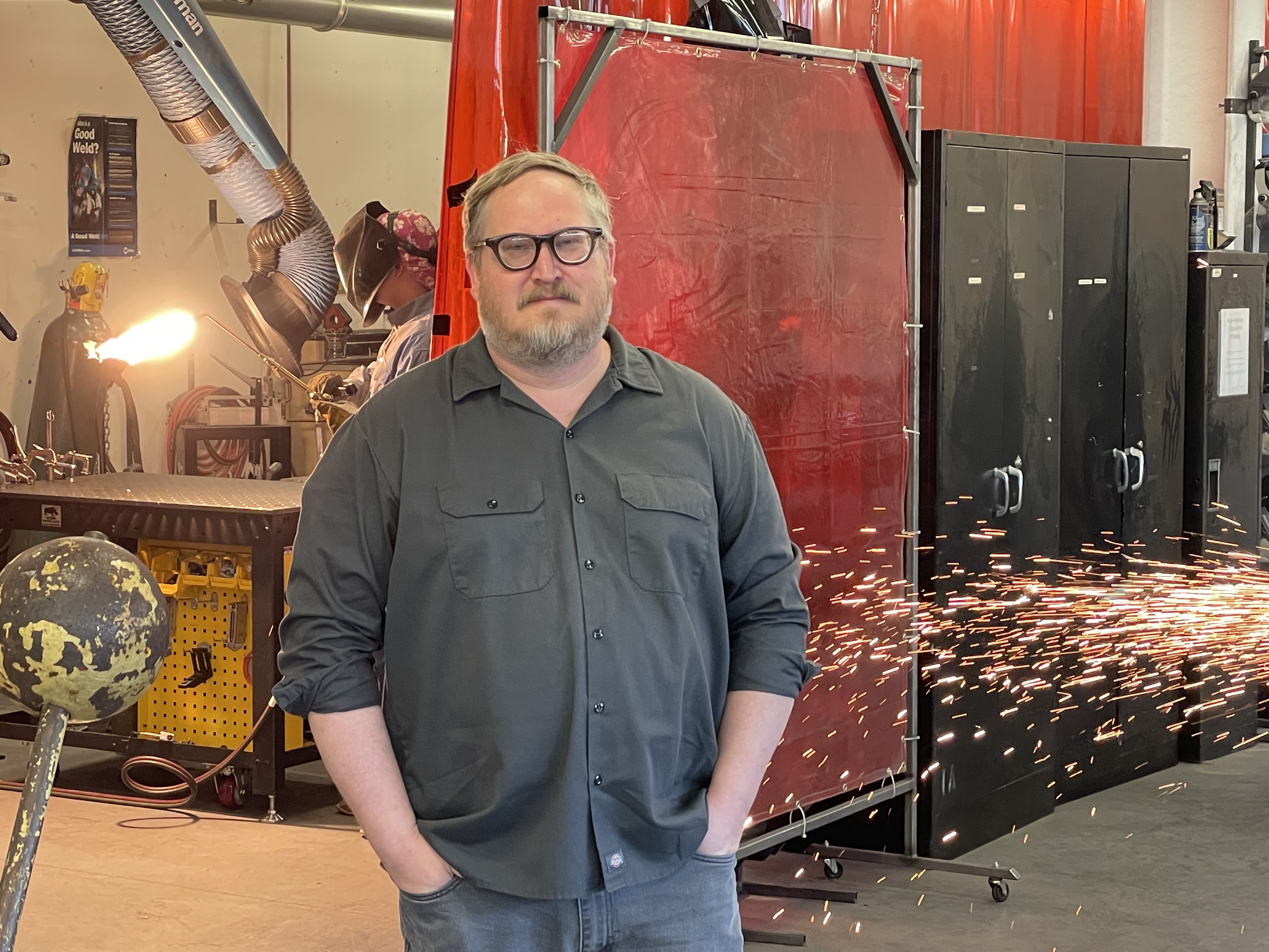 A man in a gray button-up shirt stands with his hands in his pocket in a workshop as a welder flares a torch and sparks fly behind him.