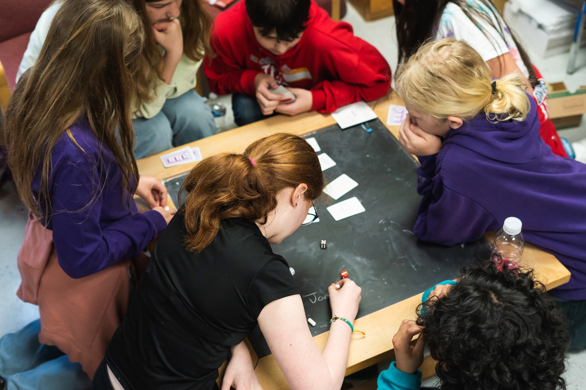 Several middle school students crowd around a chalkboard laid flat on a table and interact with it.