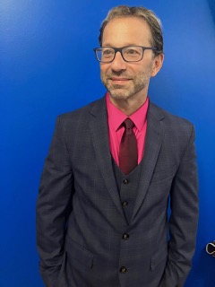 A man with hands in his pockets in a suit against a blue background.