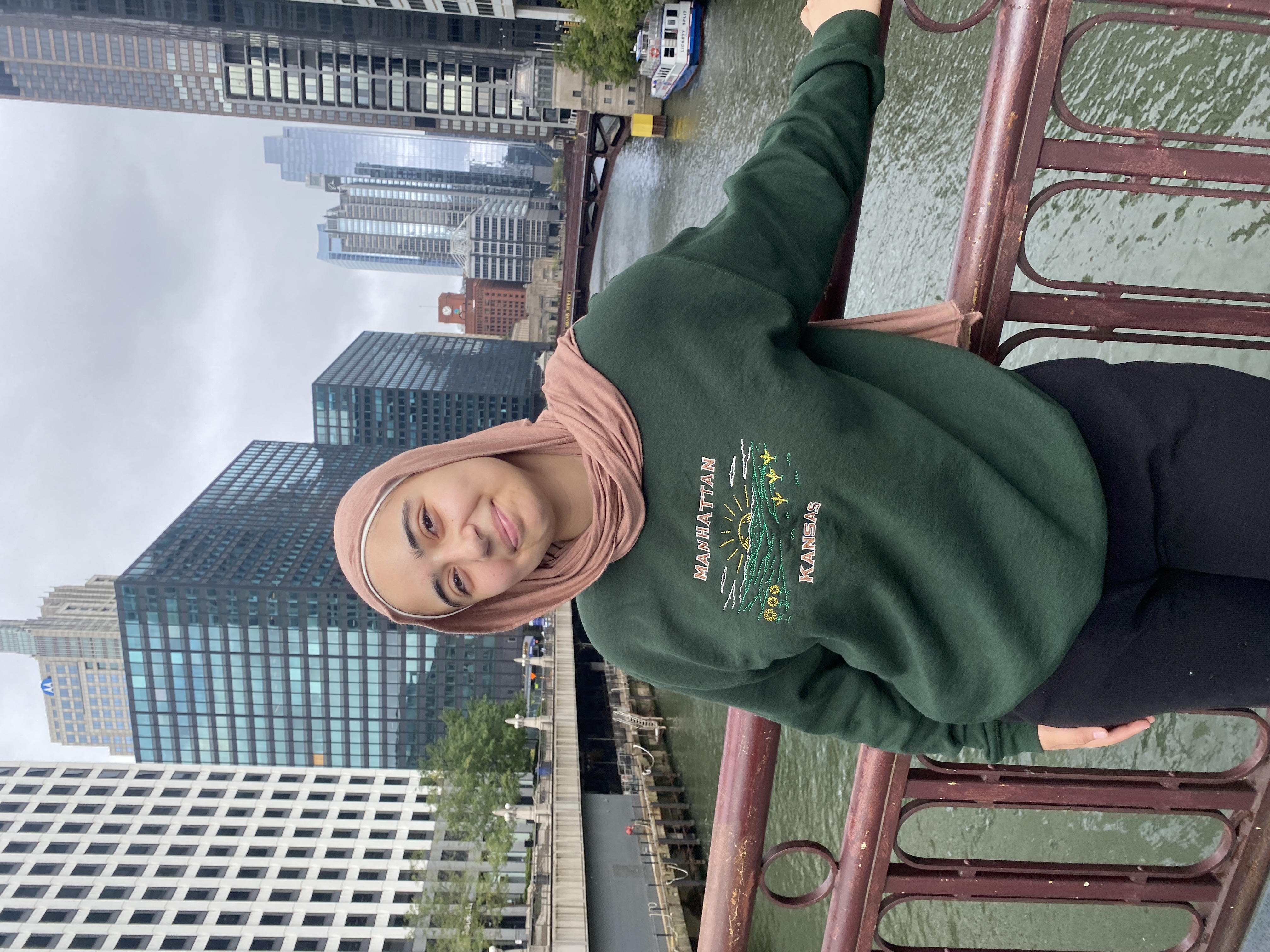 A student in a green sweater standing on a bridge overlooking a city skyline.