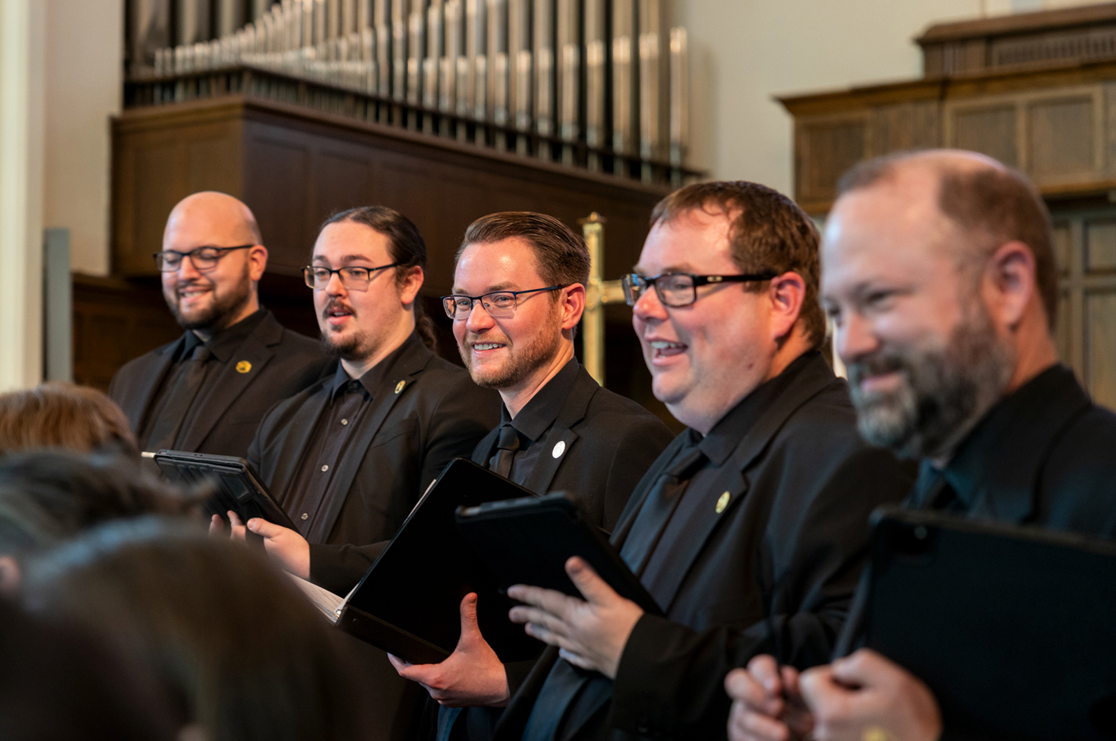 A row of five men in solid black suits, shirts and ties sing as part of the Kansas City Chorale inside a church.