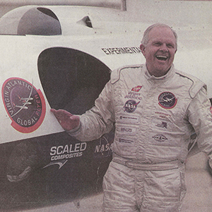 Steve Fossett, wearing a white jumpsuit, stands beside a white airplane.