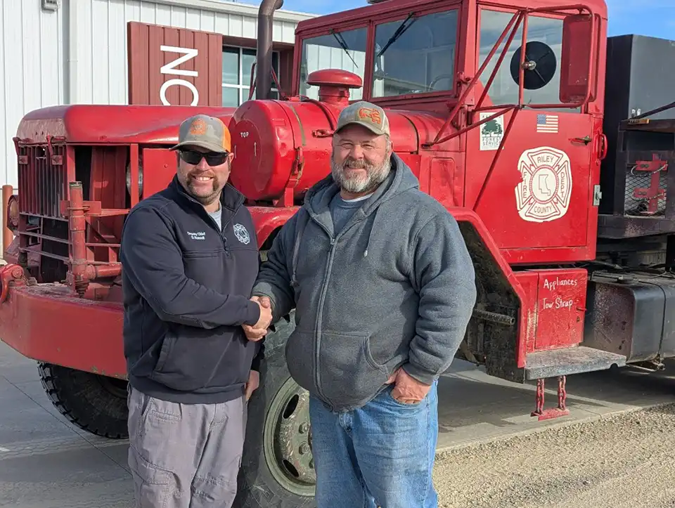 Two men shake hands in front of a red firetruck.